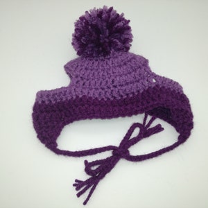 May include: A handmade, crochet hat in shades of purple. The hat features ear flaps with ties and a large pom-pom on top. The yarn appears soft and textured, suggesting a cozy, warm accessory. The hat is designed for infants or toddlers.