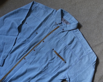 May include: A light blue long-sleeved button-down shirt with a brown trim down the front. The shirt has two chest pockets and a single pocket on the left side.