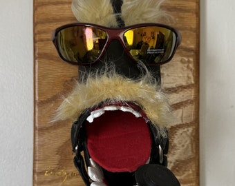 May include: A wooden plaque with a black flip-flop mounted on it. The flip-flop has a furry mustache, red mouth, and is wearing sunglasses that say "Ultraviolet Protection". The flip-flop is holding a black and white pipe.