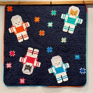May include: A dark blue quilted blanket featuring four appliqued astronauts in white suits with orange, teal, red, and blue accents. The background is decorated with various colorful star shapes and a swirling, circular stitched pattern throughout.