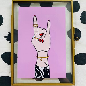 Puede incluir: Un fondo rosa con una mano haciendo el gesto de rock and roll. La mano lleva anillos de oro y una pulsera de oro con un dije de estrella. La mano tiene esmalte de u&ntilde;as rojo y un peque&ntilde;o tatuaje de coraz&oacute;n en la mu&ntilde;eca.