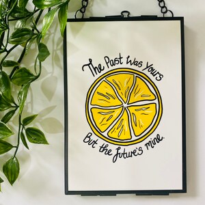 K&ouml;nnte beinhalten: Ein schwarzer gerahmter Wandbehang mit wei&szlig;em Hintergrund und einer Illustration einer gelben Zitronenscheibe. Der Text lautet "The past was yours but the future's mine."