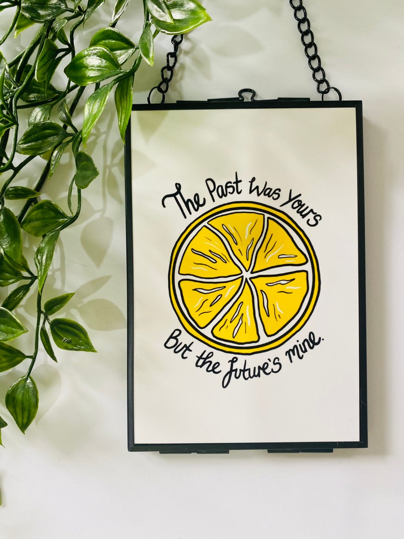 K&ouml;nnte beinhalten: Ein schwarzer gerahmter Wandbehang mit wei&szlig;em Hintergrund und einer Illustration einer gelben Zitronenscheibe. Der Text lautet "The past was yours but the future's mine."