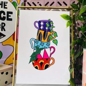 May include: A stack of four colorful mugs with different patterns, including a purple and white mug, a yellow and black mug, a blue and white mug, and an orange and black mug. The mugs are stacked on top of each other and have a green plant growing out of the top mug.
