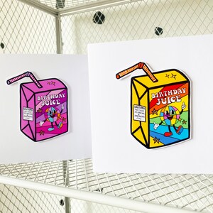 May include: Two birthday cards with a cartoon juice box design. The juice box is yellow with a rainbow design and the text "Birthday Juice". The juice box has a smiling cartoon character with a rainbow ball for a head. One card has a pink juice box and the other has a yellow juice box.