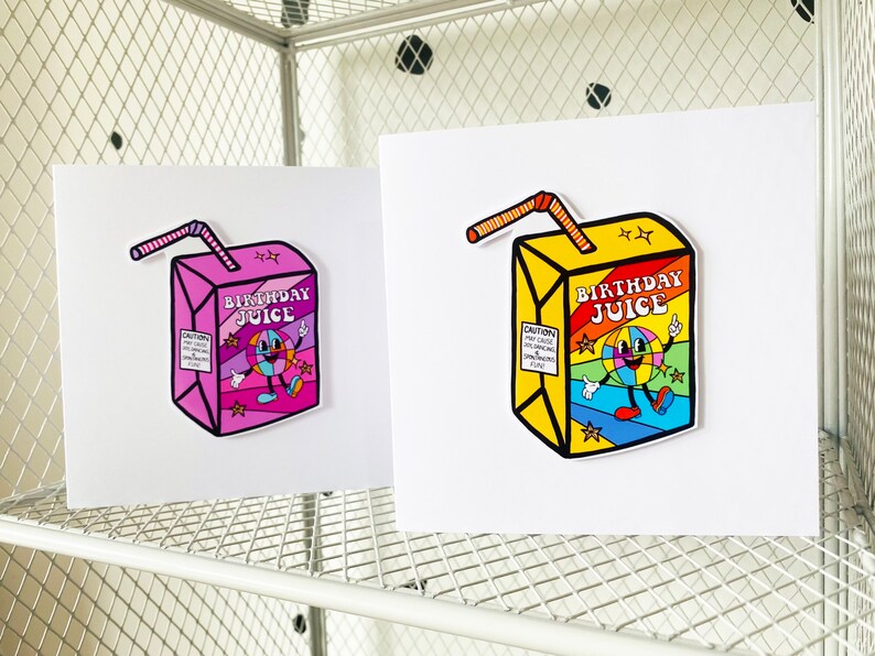 May include: Two birthday cards with a cartoon juice box design. The juice box is yellow with a rainbow design and the text "Birthday Juice". The juice box has a smiling cartoon character with a rainbow ball for a head. One card has a pink juice box and the other has a yellow juice box.