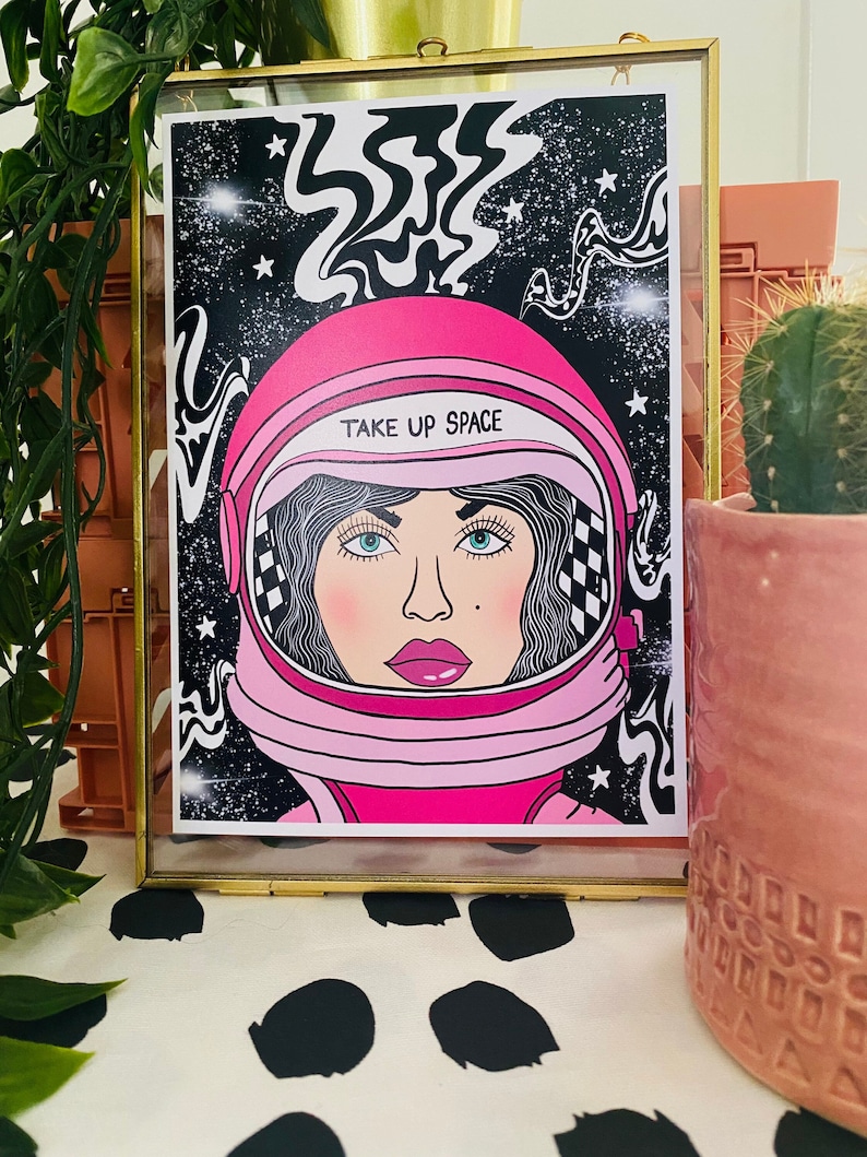 May include: A framed illustration of a woman wearing a pink astronaut helmet with the text "Take Up Space" on the visor. The background is black with white stars and swirling lines.