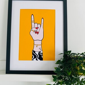 Puede incluir: Una ilustraci&oacute;n enmarcada de una mano haciendo el gesto de rock and roll. La mano lleva anillos y una pulsera. El fondo es amarillo.