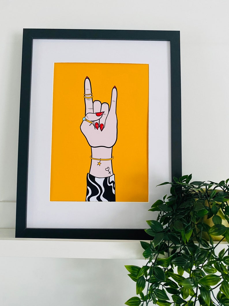 Puede incluir: Una ilustraci&oacute;n enmarcada de una mano haciendo el gesto de rock and roll. La mano lleva anillos y una pulsera. El fondo es amarillo.