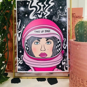 May include: A pink and white astronaut helmet with a black and white checkered pattern on the side. The helmet has the text "TAKE UP SPACE" written on it. The astronaut is set against a black background with white stars and swirling lines.