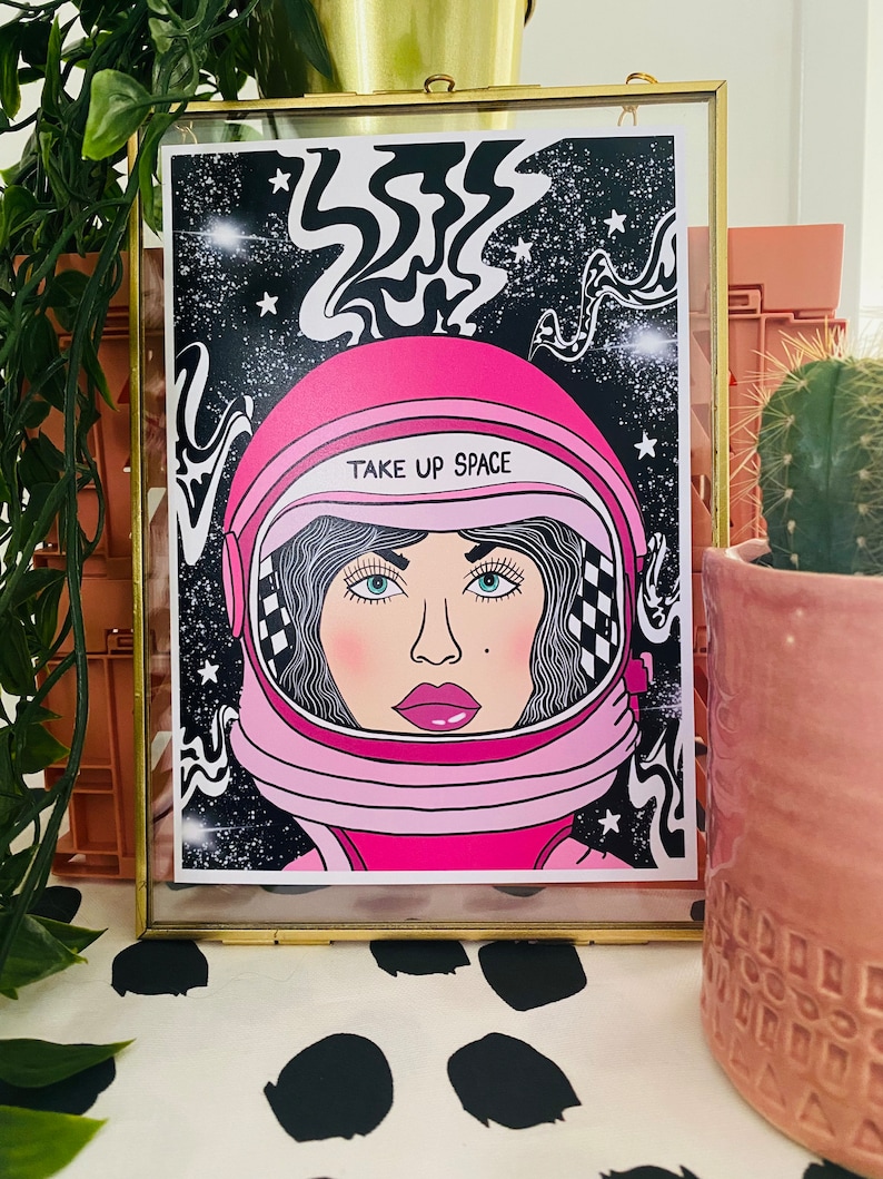 May include: A pink and white astronaut helmet with a black and white checkered pattern on the side. The helmet has the text "TAKE UP SPACE" written on it. The astronaut is set against a black background with white stars and swirling lines.
