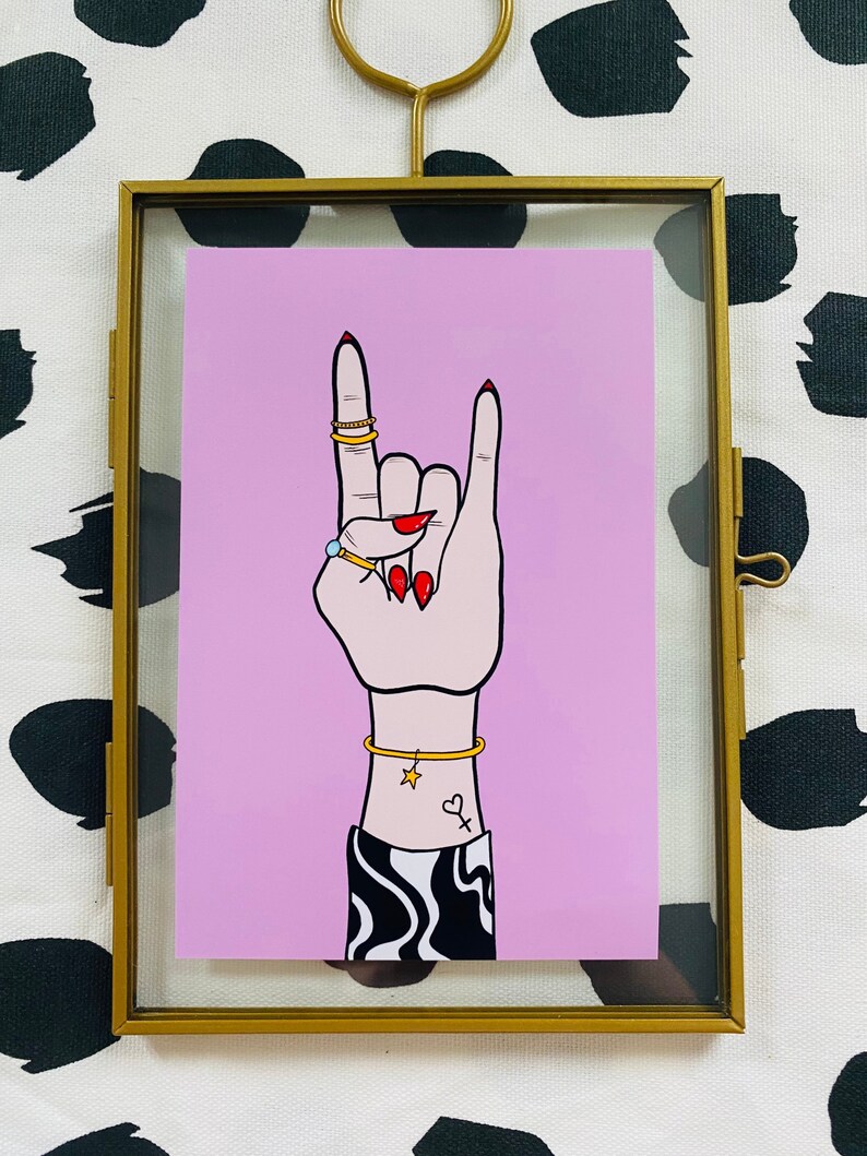 Puede incluir: Un fondo rosa con una mano haciendo el gesto de rock and roll. La mano lleva anillos de oro y una pulsera de oro con un dije de estrella. La mano tiene esmalte de u&ntilde;as rojo y un peque&ntilde;o tatuaje de coraz&oacute;n en la mu&ntilde;eca.