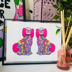 May include: A framed print of two colorful dogs with pink ears and tails. The dogs are facing each other and have a psychedelic pattern on their bodies. The print is on white paper and has a black frame.