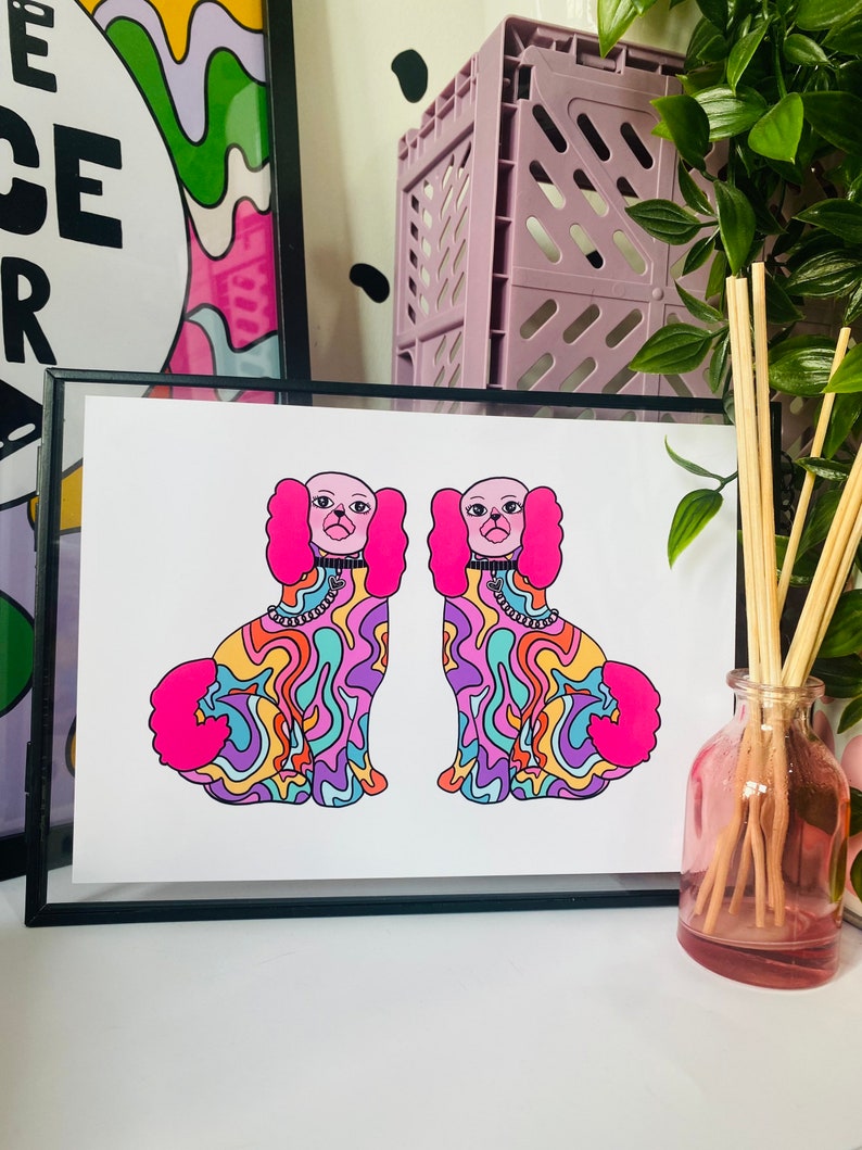 May include: A framed print of two colorful dogs with pink ears and tails. The dogs are facing each other and have a psychedelic pattern on their bodies. The print is on white paper and has a black frame.
