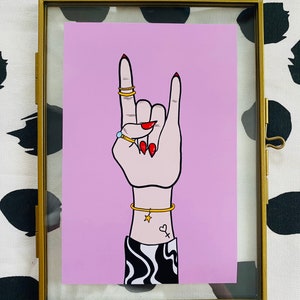 Puede incluir: Un fondo rosa con una mano haciendo el gesto de rock and roll. La mano lleva anillos de oro y una pulsera de oro. La mano tiene esmalte de u&ntilde;as rojo y un peque&ntilde;o tatuaje de estrella y coraz&oacute;n en la mu&ntilde;eca.
