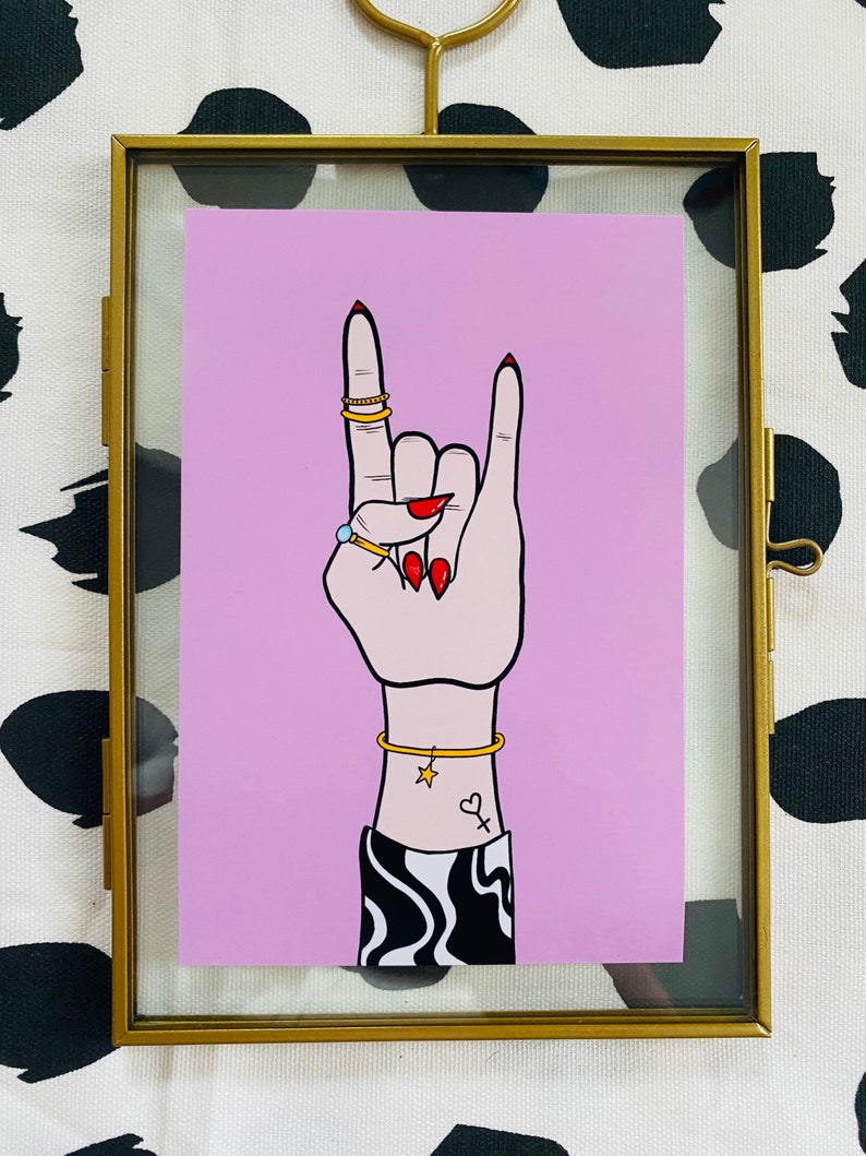 Puede incluir: Un fondo rosa con una mano haciendo el gesto de rock and roll. La mano lleva anillos de oro y una pulsera de oro. La mano tiene esmalte de u&ntilde;as rojo y un peque&ntilde;o tatuaje de estrella y coraz&oacute;n en la mu&ntilde;eca.