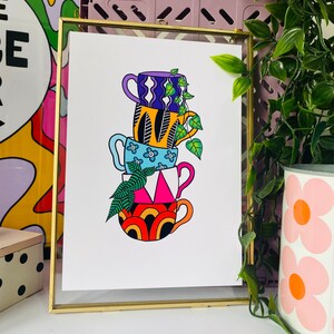 May include: A framed print of a stack of four colorful mugs with geometric patterns. The mugs are stacked on top of each other and a green plant is growing out of the top mug.