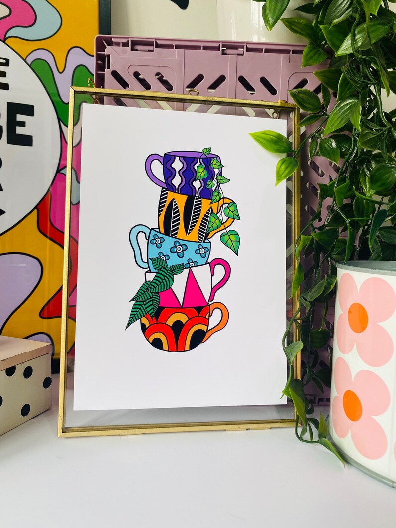 May include: A framed print of a stack of four colorful mugs with geometric patterns. The mugs are stacked on top of each other and a green plant is growing out of the top mug.