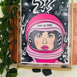 May include: A pink astronaut helmet with a black and white checkered pattern on the side. The helmet has the text "TAKE UP SPACE" written on it. The helmet is set against a black background with white stars and swirling lines.