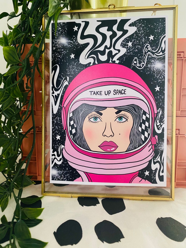 May include: A pink astronaut helmet with a black and white checkered pattern on the side. The helmet has the text "TAKE UP SPACE" written on it. The helmet is set against a black background with white stars and swirling lines.