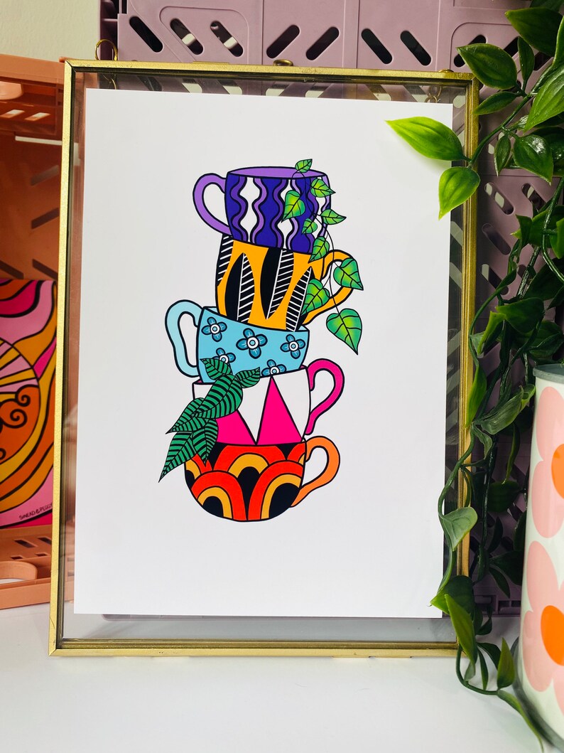 May include: A stack of four colorful mugs with geometric patterns, each with a different design. The mugs are stacked on top of each other, with a green plant growing out of the top mug. The mugs are set against a white background.