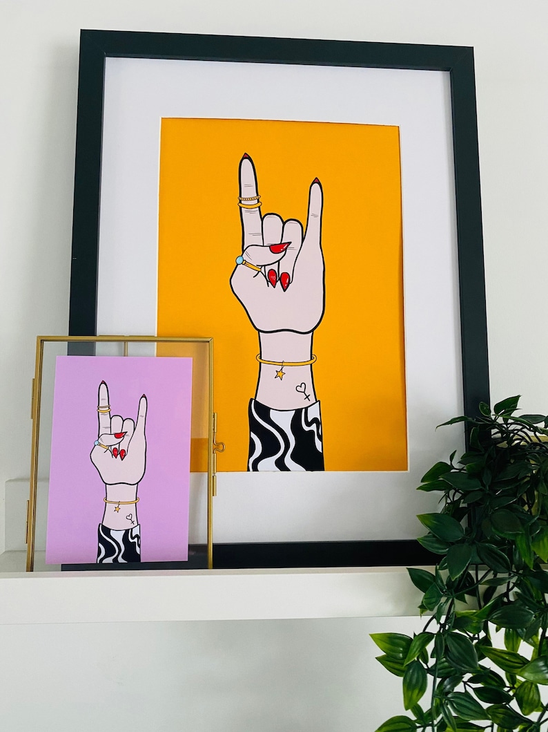 Puede incluir: Dos ilustraciones enmarcadas de una mano haciendo el gesto de rock and roll. La mano en el marco m&aacute;s grande est&aacute; sobre un fondo amarillo, mientras que la mano en el marco m&aacute;s peque&ntilde;o est&aacute; sobre un fondo rosa. Ambas manos tienen anillos y pulseras doradas, y esmalte de u&ntilde;as rojo.
