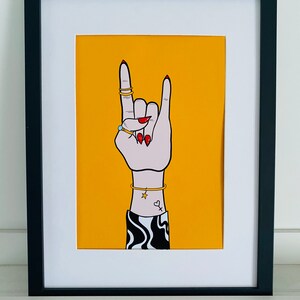 Puede incluir: Una impresi&oacute;n enmarcada en negro con un fondo amarillo que presenta una mano haciendo el gesto de rock and roll. La mano lleva anillos y una pulsera con un dije de estrella. La mano tiene esmalte de u&ntilde;as rojo y un peque&ntilde;o tatuaje de coraz&oacute;n.