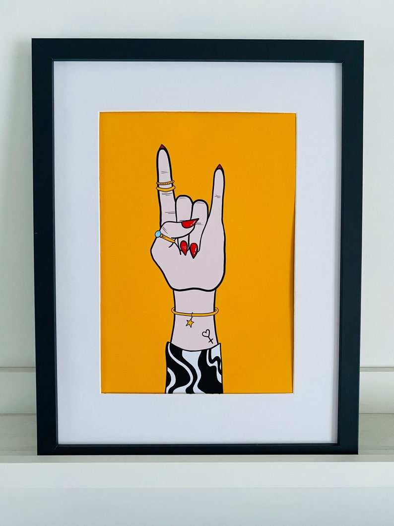 Puede incluir: Una impresi&oacute;n enmarcada en negro con un fondo amarillo que presenta una mano haciendo el gesto de rock and roll. La mano lleva anillos y una pulsera con un dije de estrella. La mano tiene esmalte de u&ntilde;as rojo y un peque&ntilde;o tatuaje de coraz&oacute;n.