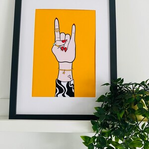 Puede incluir: Una impresi&oacute;n enmarcada de una mano haciendo el gesto de rock and roll. La mano lleva anillos y pulseras y tiene un peque&ntilde;o tatuaje de coraz&oacute;n. El fondo es de color amarillo brillante.