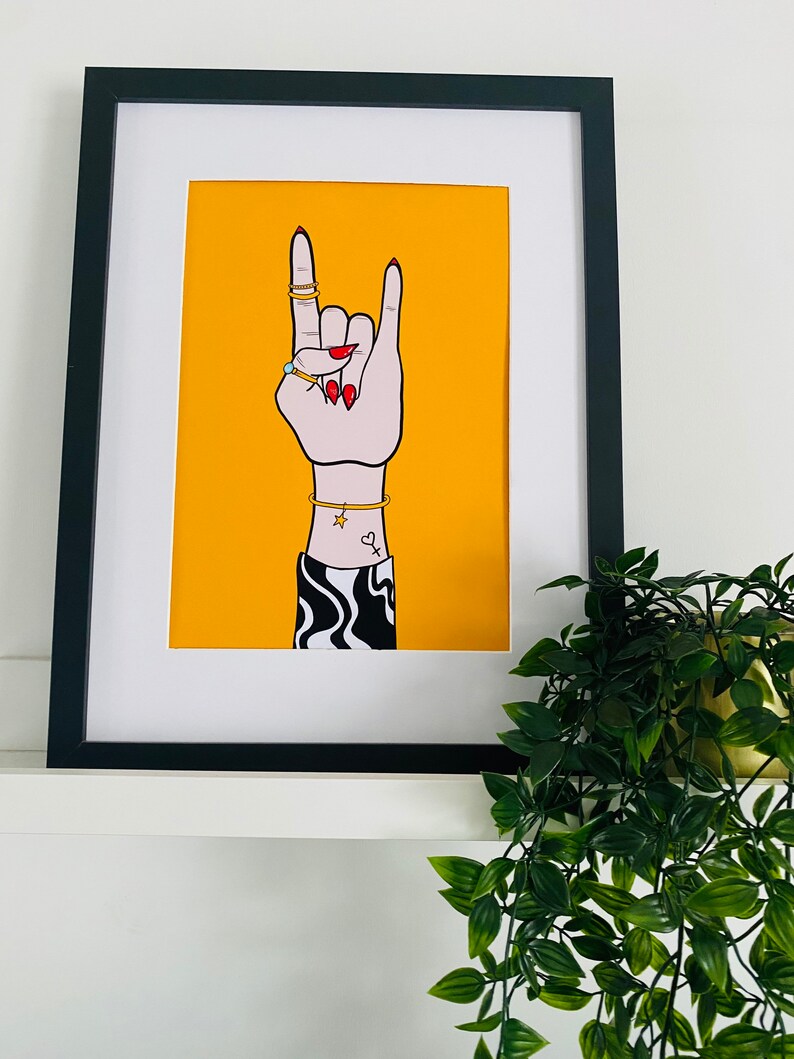 Puede incluir: Una impresi&oacute;n enmarcada de una mano haciendo el gesto de rock and roll. La mano lleva anillos y pulseras y tiene un peque&ntilde;o tatuaje de coraz&oacute;n. El fondo es de color amarillo brillante.