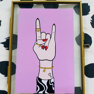 Puede incluir: Un fondo rosa con una mano haciendo el gesto de rock and roll. La mano lleva anillos de oro y una pulsera de oro con un dije de estrella. La mano tiene esmalte de u&ntilde;as rojo y un tatuaje de un coraz&oacute;n y un s&iacute;mbolo en la mu&ntilde;eca.