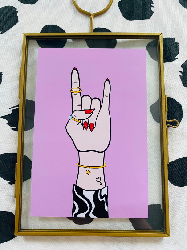 Puede incluir: Un fondo rosa con una mano haciendo el gesto de rock and roll. La mano lleva anillos de oro y una pulsera de oro con un dije de estrella. La mano tiene esmalte de u&ntilde;as rojo y un tatuaje de un coraz&oacute;n y un s&iacute;mbolo en la mu&ntilde;eca.