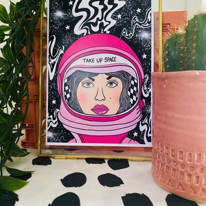 May include: A framed illustration of a woman wearing a pink astronaut helmet with the text "TAKE UP SPACE" on it. The background is black with white stars and swirls.