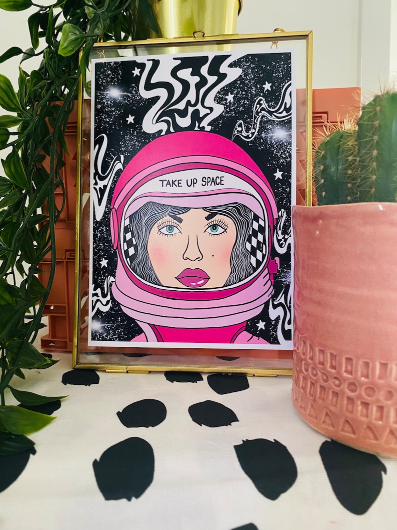 May include: A framed illustration of a woman wearing a pink astronaut helmet with the text "TAKE UP SPACE" on it. The background is black with white stars and swirls.