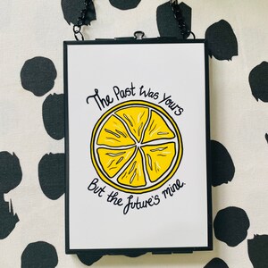 K&ouml;nnte beinhalten: Ein schwarzer gerahmter Wandbehang mit wei&szlig;em Hintergrund und einer gelben Zitronenscheibengrafik. Der Text "The Past Was Yours But the Future's Mine" ist in schwarzer Kursivschrift geschrieben.
