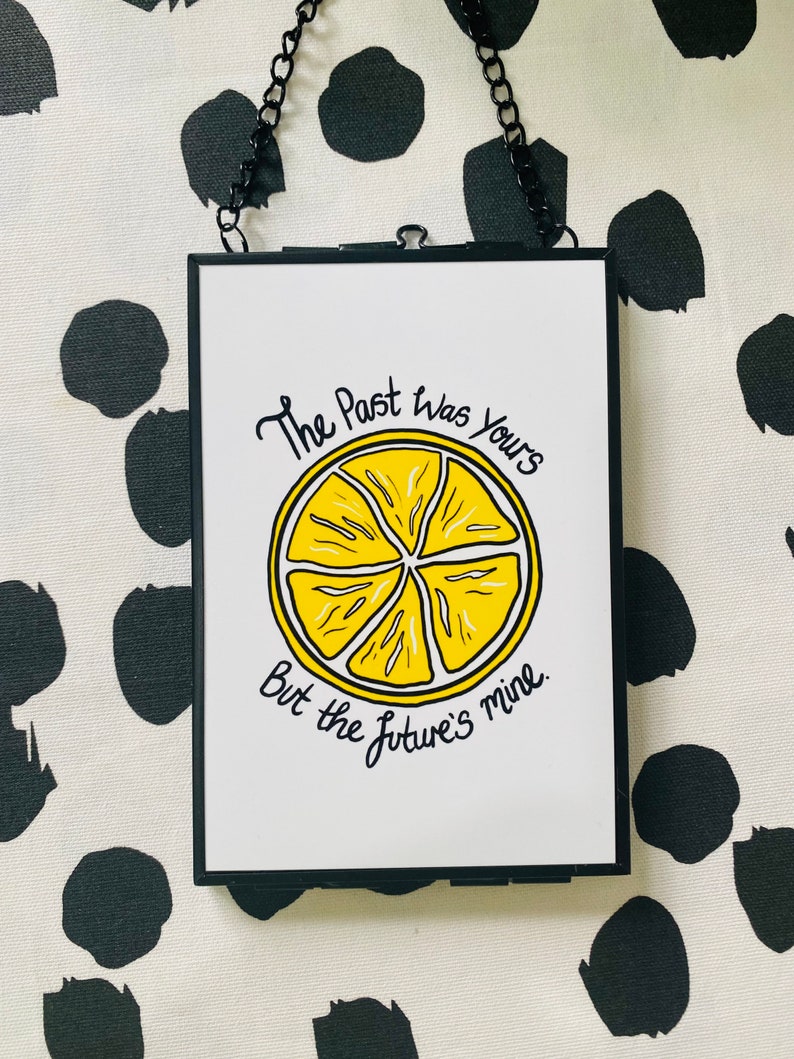 K&ouml;nnte beinhalten: Ein schwarzer gerahmter Wandbehang mit wei&szlig;em Hintergrund und einer gelben Zitronenscheibengrafik. Der Text "The Past Was Yours But the Future's Mine" ist in schwarzer Kursivschrift geschrieben.
