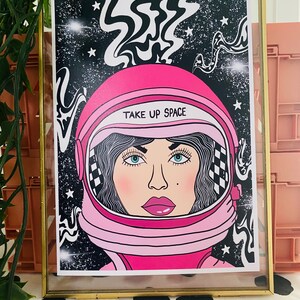 May include: A pink astronaut helmet with a black and white swirl design in the background. The helmet has a white visor and the text "TAKE UP SPACE" on the front.