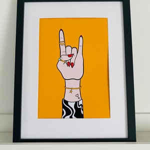 Puede incluir: Un fondo amarillo con una mano blanca haciendo el gesto de rock and roll. La mano lleva anillos dorados y tiene esmalte de u&ntilde;as rojo. La mano tiene un peque&ntilde;o tatuaje de estrella y coraz&oacute;n en la mu&ntilde;eca. La mano lleva una manga con estampado blanco y negro.