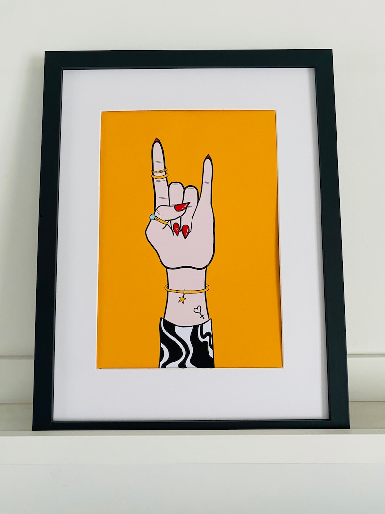 Puede incluir: Un fondo amarillo con una mano blanca haciendo el gesto de rock and roll. La mano lleva anillos dorados y tiene esmalte de u&ntilde;as rojo. La mano tiene un peque&ntilde;o tatuaje de estrella y coraz&oacute;n en la mu&ntilde;eca. La mano lleva una manga con estampado blanco y negro.