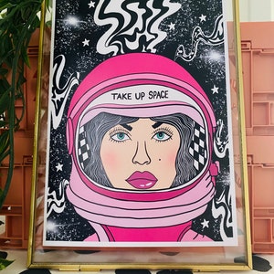 May include: A pink astronaut helmet with a woman's face inside. The helmet has a black and white checkered pattern on the side and the text "TAKE UP SPACE" on the front. The background is black with white stars and swirling lines.