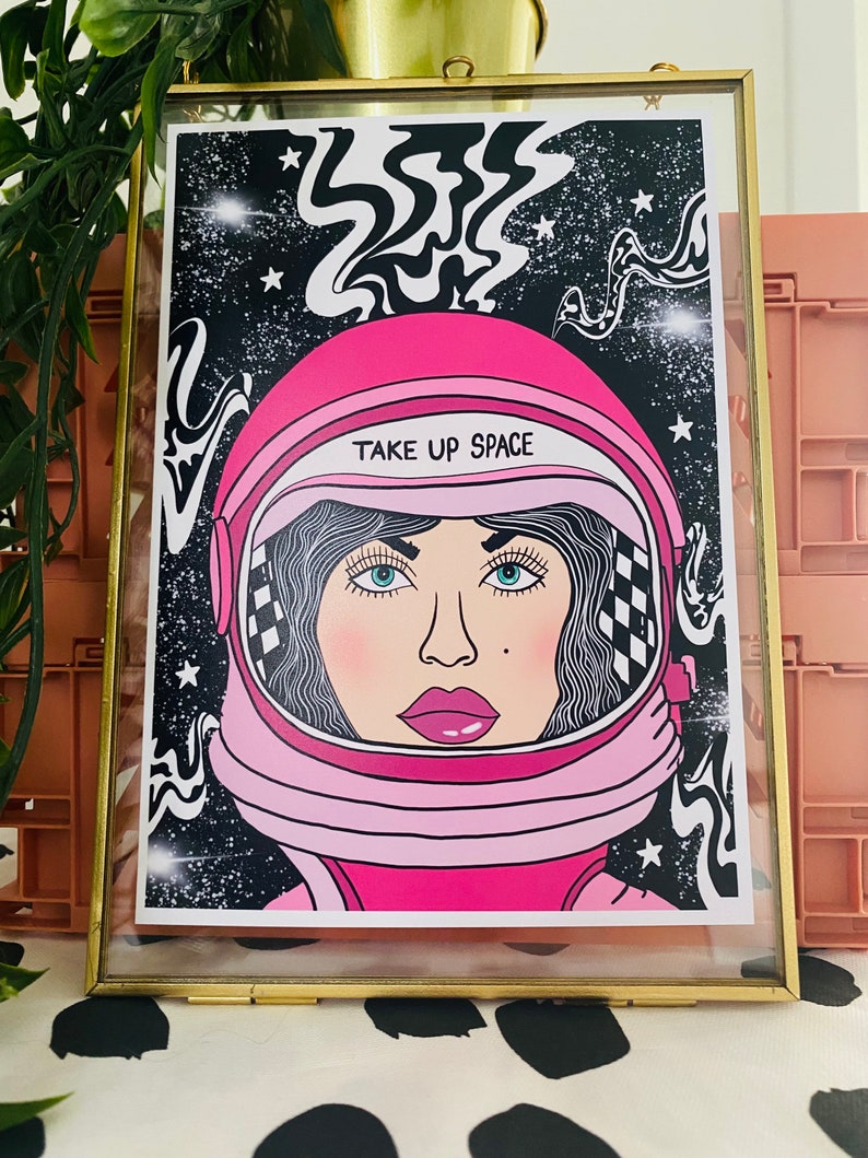 May include: A pink astronaut helmet with a woman's face inside. The helmet has a black and white checkered pattern on the side and the text "TAKE UP SPACE" on the front. The background is black with white stars and swirling lines.