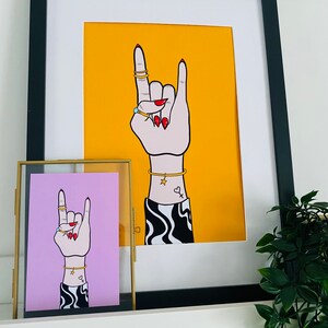 Puede incluir: Dos ilustraciones enmarcadas de una mano haciendo el gesto de rock and roll. La mano lleva anillos y pulseras. La ilustraci&oacute;n de la izquierda est&aacute; enmarcada en oro y tiene un fondo rosa. La ilustraci&oacute;n de la derecha est&aacute; enmarcada en negro y tiene un fondo amarillo.