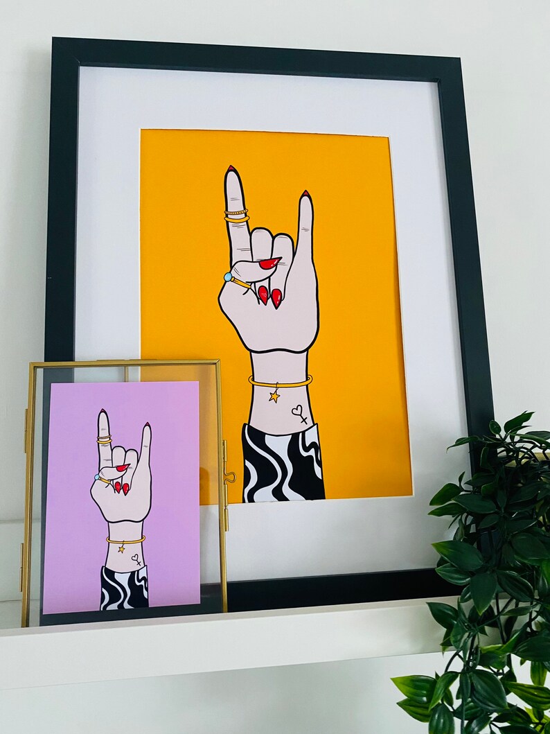Puede incluir: Dos ilustraciones enmarcadas de una mano haciendo el gesto de rock and roll. La mano lleva anillos y pulseras. La ilustraci&oacute;n de la izquierda est&aacute; enmarcada en oro y tiene un fondo rosa. La ilustraci&oacute;n de la derecha est&aacute; enmarcada en negro y tiene un fondo amarillo.