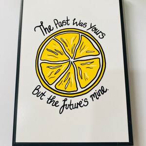 K&ouml;nnte beinhalten: Ein schwarzer gerahmter Wandbehang mit wei&szlig;em Hintergrund und einer gelben Zitronenscheibengrafik. Der Text "The Past was Yours But the Future's Mine" ist in schwarzer Kursivschrift geschrieben.