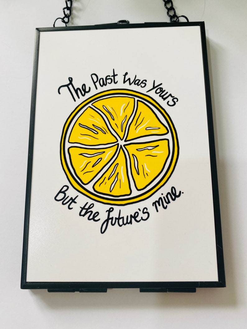 K&ouml;nnte beinhalten: Ein schwarzer gerahmter Wandbehang mit wei&szlig;em Hintergrund und einer gelben Zitronenscheibengrafik. Der Text "The Past was Yours But the Future's Mine" ist in schwarzer Kursivschrift geschrieben.