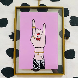 Puede incluir: Una ilustraci&oacute;n enmarcada de una mano haciendo el gesto de rock and roll. La mano lleva anillos de oro y una pulsera de oro. La mano est&aacute; sobre un fondo rosa con l&iacute;neas onduladas en blanco y negro en la manga.