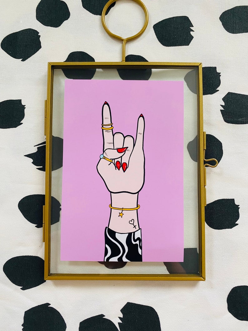 Puede incluir: Una ilustraci&oacute;n enmarcada de una mano haciendo el gesto de rock and roll. La mano lleva anillos de oro y una pulsera de oro. La mano est&aacute; sobre un fondo rosa con l&iacute;neas onduladas en blanco y negro en la manga.