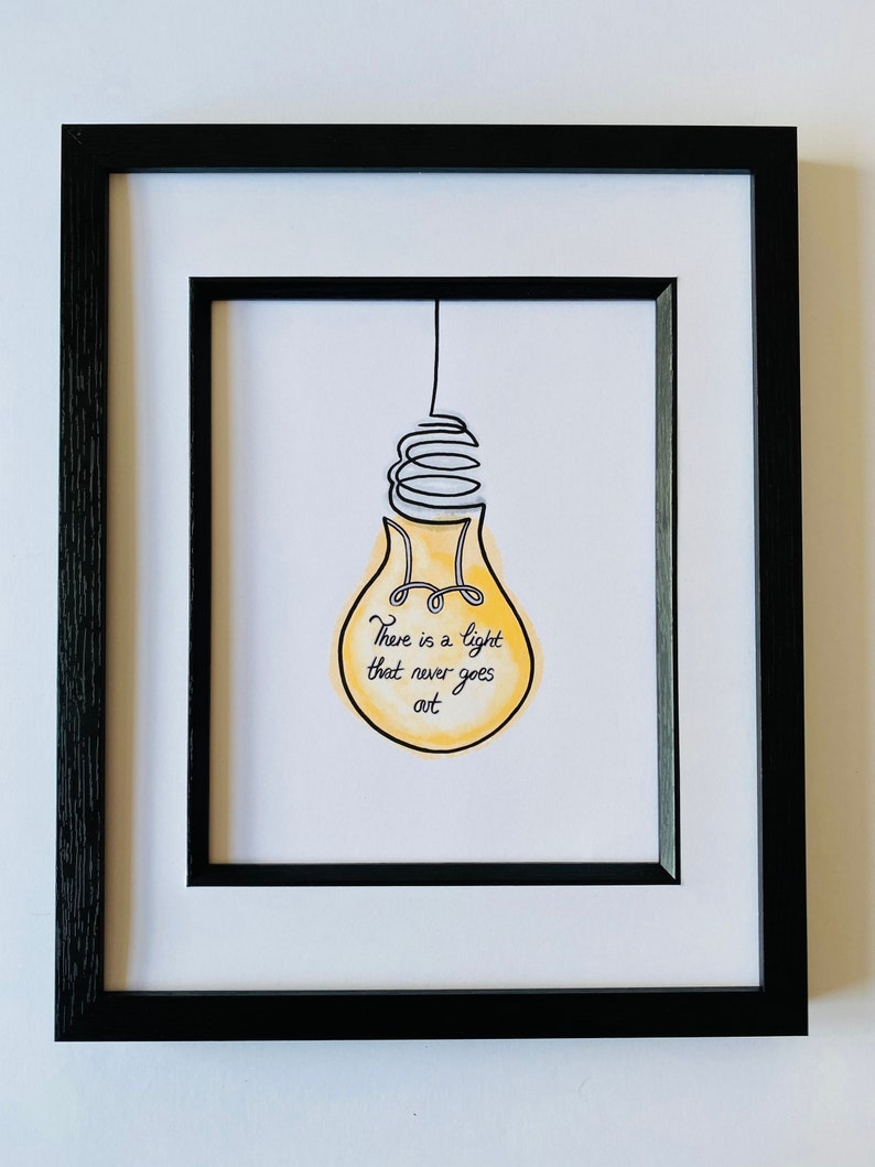 May include: A black framed print with a white mat. The print features a yellow light bulb with a black wire and the text "There is a light that never goes out."