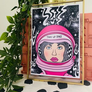 May include: A framed print with a pink astronaut helmet and a woman's face inside. The background is black with white stars and swirls. The text "TAKE UP SPACE" is written on the helmet.