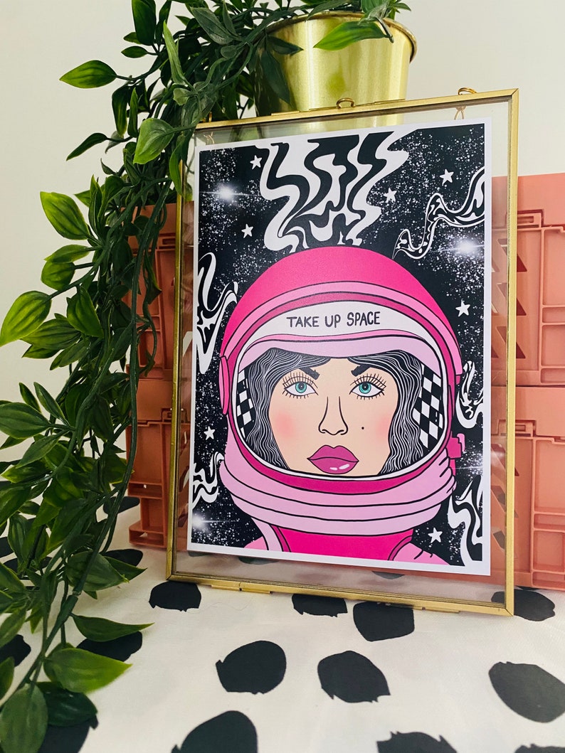 May include: A framed print with a pink astronaut helmet and a woman's face inside. The background is black with white stars and swirls. The text "TAKE UP SPACE" is written on the helmet.